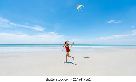 Smiling little Asian child girl in swimwear running and playing kite on tropical beach in summer sunny day. Happy children enjoy and fun outdoor activity lifestyle on travel beach vacation at the sea