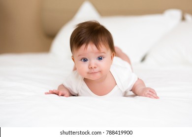 Smiling little baby girl with big blue eyes lying on her tummy on white bed looking at camera and smiling