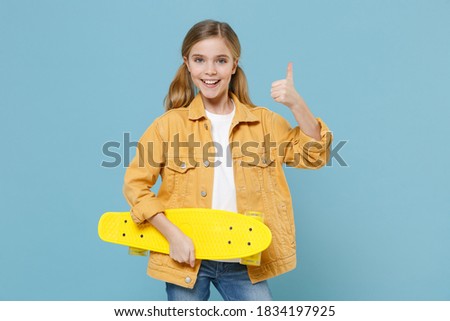 Smiling little blonde kid girl 12-13 years old in casual yellow jacket posing isolated on blue background studio. Childhood lifestyle concept. Mock up copy space. Hold skateboard, showing thumb up