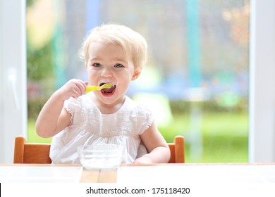 Smiling little blonde toddler girl eating delicious yogurt sitting in the kitchen in a high feeding chair next to a big window with garden view