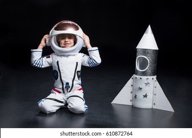 Smiling Little Boy Astronaut In Space Suit Kneeling Near Rocket And Adjusting Helmet