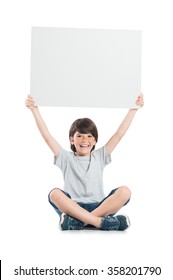 Smiling Little Boy Holding Placard Isolated On White Background. Happy Cute Boy Holding Blank Poster Sitting On White Floor. Laughing Child Showing Empty White Sign.