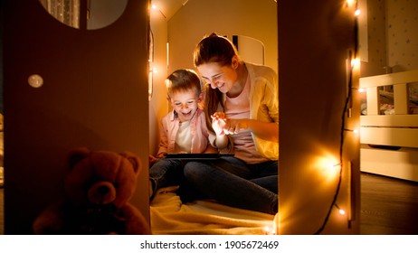 Smiling Little Boy With Mother Playing In Toy Cardboard House And Using Tablet Computer. Concept Of Child Education And Family Having Time Together At Night.