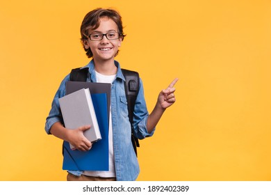 Smiling little boy pointing at copy space in casual clothes with books for studing at school isolated over yellow background