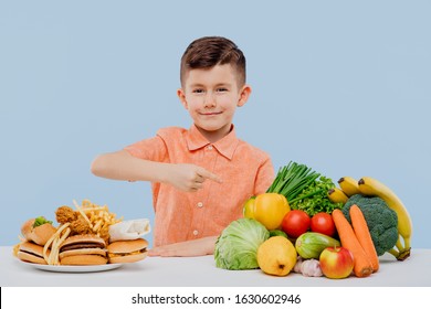 Smiling Little Boy Pointing At Healthy Food. Fruits, Vegetables And Junk Food. Healthy Food And Fast Food Concept, Isolated On Blue Background. Studio. At The Table