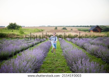 Smiling little boy running on the lavender field. Funny child in glasses dressed in a blue shirt and a straw hat running on a grass between lavender flowers. Horizontal photo