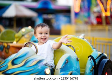 Smiling Little Boy Sitting In A Tiny Car On A Merry Go Round (carousel) In The Theme Park And Pointing At Something. Leisure With Kids Concept. Room For Copy Text