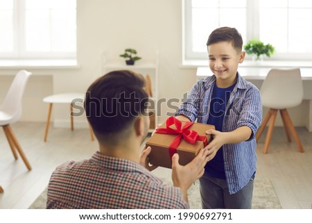 Smiling little boy son congratulating giving to dad wrapped gift box surprising with present greeting with father day or birthday. Home living-room interior. Happy family and holidays celebration