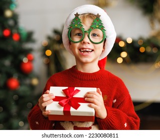 Smiling little boy wearing funny glasses in form of Christmas trees    holding Xmas gift while standing  in a decorated holiday room at home, dressed in warm red xmas sweater. 