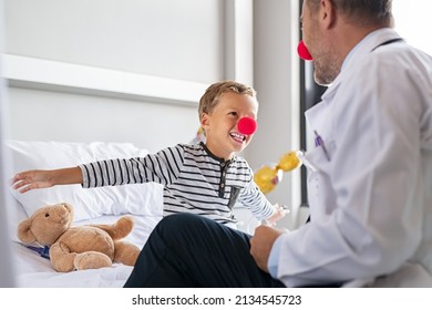 Smiling little boy wearing red nose and flying with outstretched arms like superhero in hospital bed with doctor. Kid joking during medical examination with physician, clown therapy concept. 