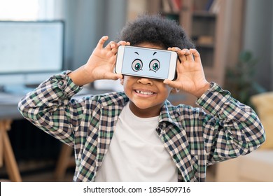 Smiling little boy in white t-shirt and checkered shirt holding smartphone with funny eyes on screen in front of his face against home environment