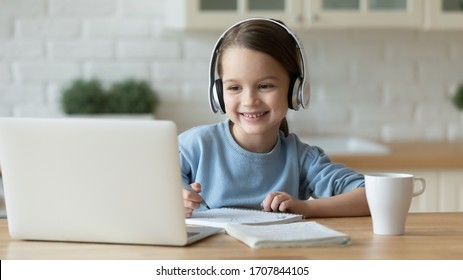 Smiling Little Caucasian Girl In Headphones Handwrite Study Online Using Laptop At Home, Cute Happy Small Child In Earphones Take Internet Web Lesson Or Class On Computer, Homeschooling Concept