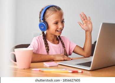 Smiling Little Caucasian Girl In Headphones Having Video Call Distant Class With Teacher While Using Laptop. Happy Small Child Waving And Greeting With Tutor. Study Online On Computer, Homeschooling