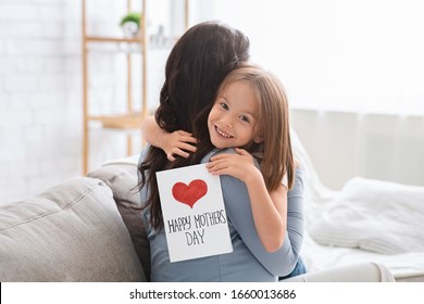 Smiling Little Daughter With Happy Mother's Day Gift Card Hugging Her Mom, Celebrating International Mother's Day Together At Home