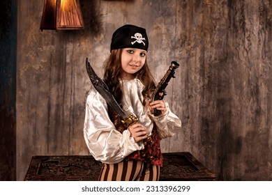 Smiling little girl actress 7-8 year old in pirate's image with gun and saber at gray textured wall, looking at camera. Kid in pirate corsair costume on stage. Theatre style concept. Copy text space
