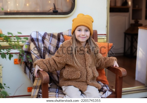 Smiling little girl in casual clothes and hat\
sitting on chair on porch at house. Child in cozy RV campsite fall\
backyard. Concept camping, adventure. Girl resting and walking in\
autumn garden
