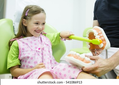 Smiling little girl in dentists chair, being educated about proper tooth-brushing by her paediatric dentist. Early prevention, raising awareness, oral hygiene demonstration concept. 
