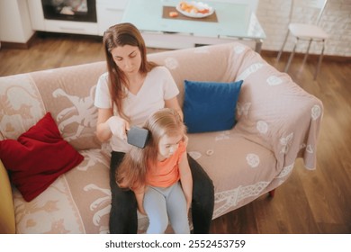 A smiling little girl enjoys her mother combing her long hair on a comfortable sofa, highlighting love, care, and family bonding in a warm indoor setting. - Powered by Shutterstock