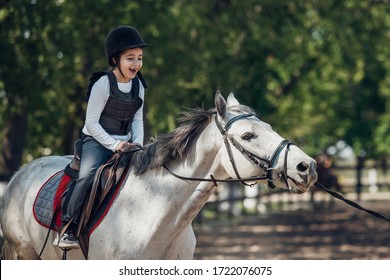 Smiling Little Girl In Helmet Learning Horseback Riding. Instructor Teaches Kid Equestrian.