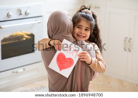 Smiling little girl holding greeting card for Happy Mother's Day with drawn red heart and hugging her muslim mom. Loving islamic family bonding together at home, closeup shot with free space