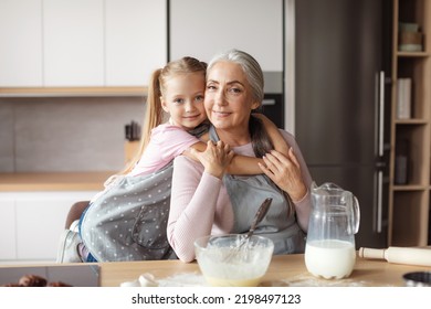 Smiling Little Girl Hugs Old Woman In Apron Making Dough In Kitchen Interior. Household Chores Together, Cooking Lesson Baking, Prepare Food At Home. Granddaughter And Grandmother Relationship, Love