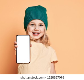 Smiling Little Girl Kid In Green Modern Winter Hat Showing White Blank Screen For Text Space Of New Popular Mobile Phone On Light Yellow Background