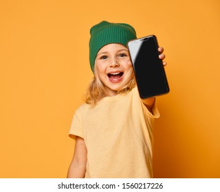 Smiling Little Girl Kid In Green Modern Winter Hat Showing Blank Screen Of New Popular Mobile Phone On Light Yellow Background