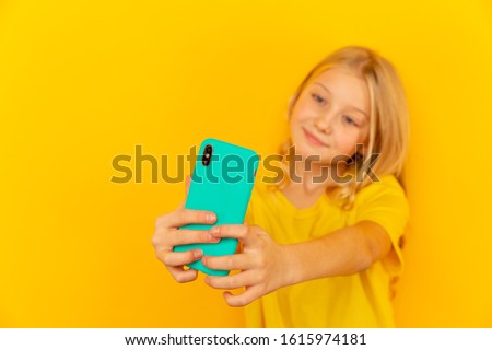 Smiling little girl kid showing blue screen of new popular mobile phone on light yellow background.