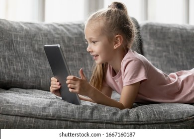 Smiling little girl lying on comfortable couch, enjoying playing online game on digital tablet computer. Addicted to technology, happy small kid using funny applications, web surfing information.