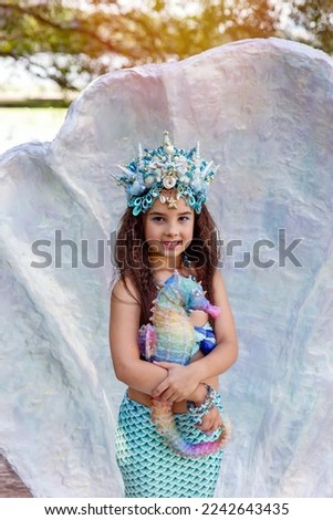 A smiling little girl in a mermaid costume in a large seashell, holding a colorful seahorse toy.