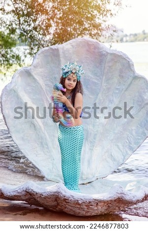 A smiling little girl in a mermaid costume in a large seashell, holding a colorful seahorse toy.