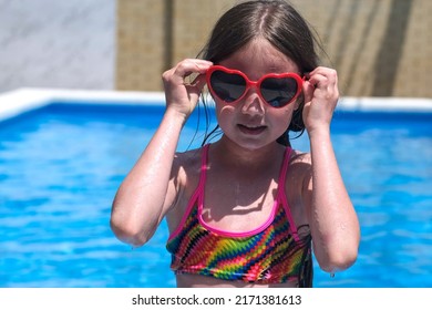 Smiling Little Girl On Sunny Day Puts On Heart Shaped Sunglasses On Background Of Swimming Pool. Childhood, Valentines Day And Summer Vacation Concept. Happy Kid Having Fun Outdoors On Holidays