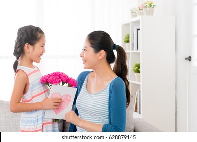 Smiling Little Girl Prepare A Pink Carnation Bouquet And Love Card Giving Gift For Beautiful Mom At Mother's Day At Home In The Living Room Sofa Looking Each Other.