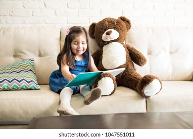 Smiling little girl sitting on couch and reading fairy tale book with teddy bear in the living room at home.