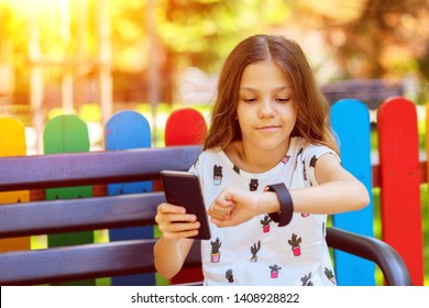 Smiling Little Girl Using Modern Wearable Smart Watch And Mobile Phone Outdoor Synching Data – Happy Kid With Trendy Gadgets Synchronizing Smartwatch With Smartphone While Sitting On Bench In Park