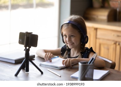 Smiling Little Girl Wearing Headphones Studying Online At Home, Looking At Phone Screen, Watching Webinar, Engaged In Video Conference, Using Smartphone On Tripod, Writing Notes, Homeschooling