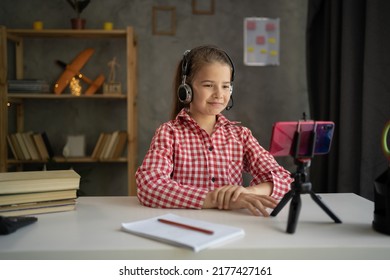 Smiling Little Girl Wearing Headphones Studying Online At Home, Looking At Phone Screen, Engaged In Video Conference, Watching Webinar, Using Smartphone On Tripod, Homeschooling Concept