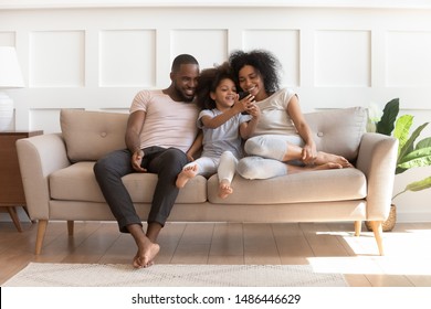 Smiling little mixed race girl holding smartphone, playing game, showing funny video to loving parents. Happy african american family sitting on comfortable couch in modern living room, using phone. - Powered by Shutterstock