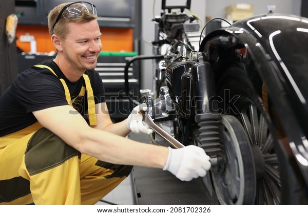Smiling locksmith\
with wrench near motorcycle wheel in car workshop. Motorcycle\
warranty service\
concept