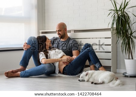 Smiling love couple expecting baby enjoying in their empty apartment with their cute white dog. How to Introduce Dogs and Babies