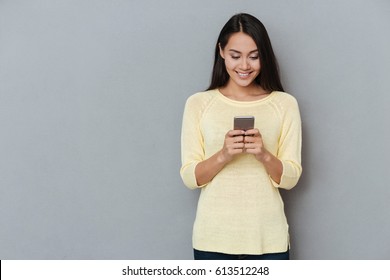 Smiling lovely young woman standing and using cell phone over grey background