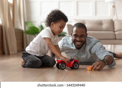 Smiling loving african American dad lying on floor playing with toddler son with toy cars at home, happy caring black father have fun with little kid engaged in funny activity race on automobiles