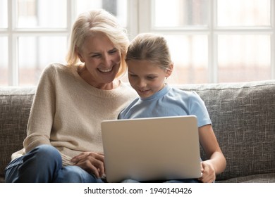 Smiling loving mature grandmother and small teen granddaughter relax on couch at home use modern computer gadget together. Happy older granny and little girl child have fun browsing web on laptop. - Powered by Shutterstock