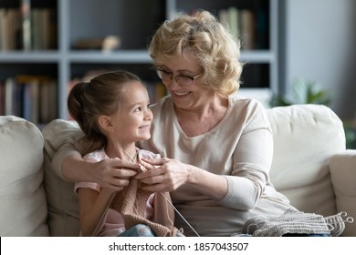 Smiling loving mature grandmother wearing glasses teaching little granddaughter knitting, hugging, sitting on couch at home, positive middle aged woman and cute girl enjoying leisure time