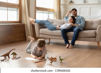 Smiling loving young bonding couple parents relaxing on comfortable sofa, watching little kid son drawing pictures in album, lying on wooden floor in living room, happy family weekend pastime.