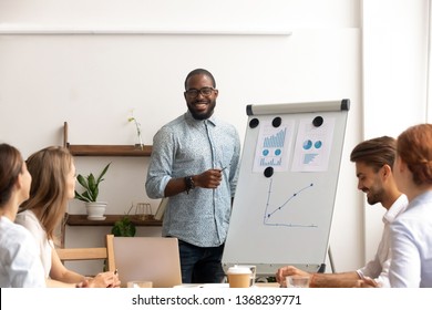 Smiling Male African Conference Speaker Give Flipchart Presentation Explaining Work Result Training Sales Team, Black Coach Trainer Mentor Consulting Clients Interns At Corporate Group Office Seminar