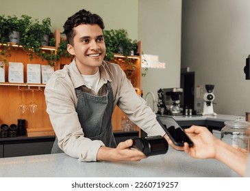 Smiling male barista holding pos terminal looking at the customer. Bartender receive contactless payment.