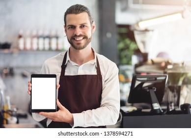 Smiling Male Barista Holding And Showing Digital Tablet With White Blank Screen At The Coffee Shop Or Restaurant Cafe. Beard Man With Startup Of Small Business Entrepreneur Owner. Looking Camera.