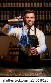 Smiling Male Bartender In Black Apron Skillfully Pours A Steaming Cocktail From One Shaker Cup To Another. Blue Backlit