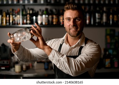 Smiling Male Bartender In Black Leather Apron Holding A Glass Shaker In His Hands. Blurred Shelves With Bottles In The Background